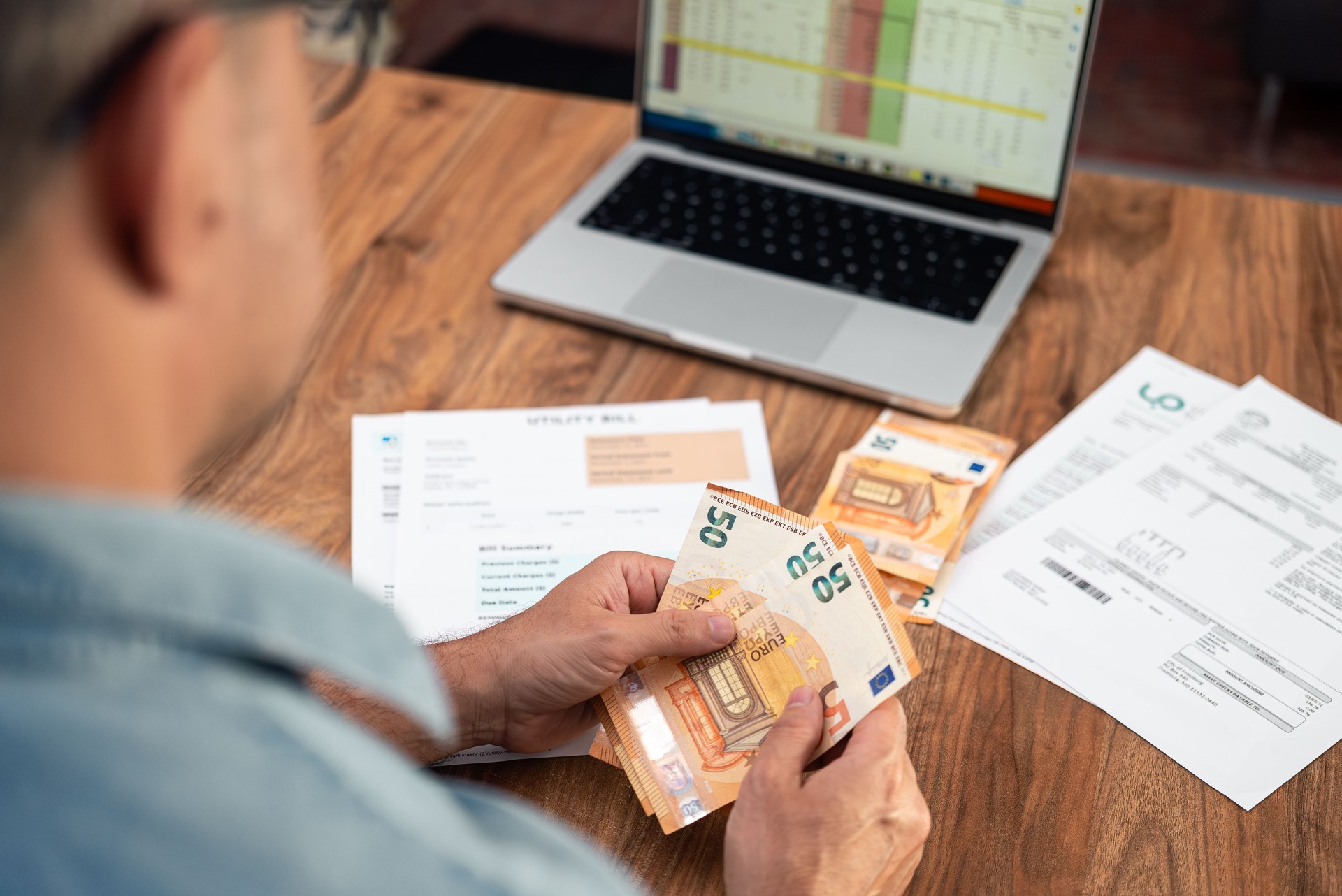Old man counts euros banknotes sitting at table with unpaid utility bills and laptop in office